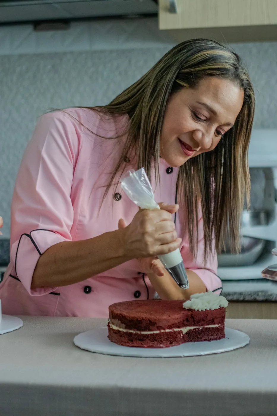 Woman in a pink chef jacket skillfully decorates a cake with frosting in a kitchen setting. Licensed inspiration image; not a BMG client portfolio claim.